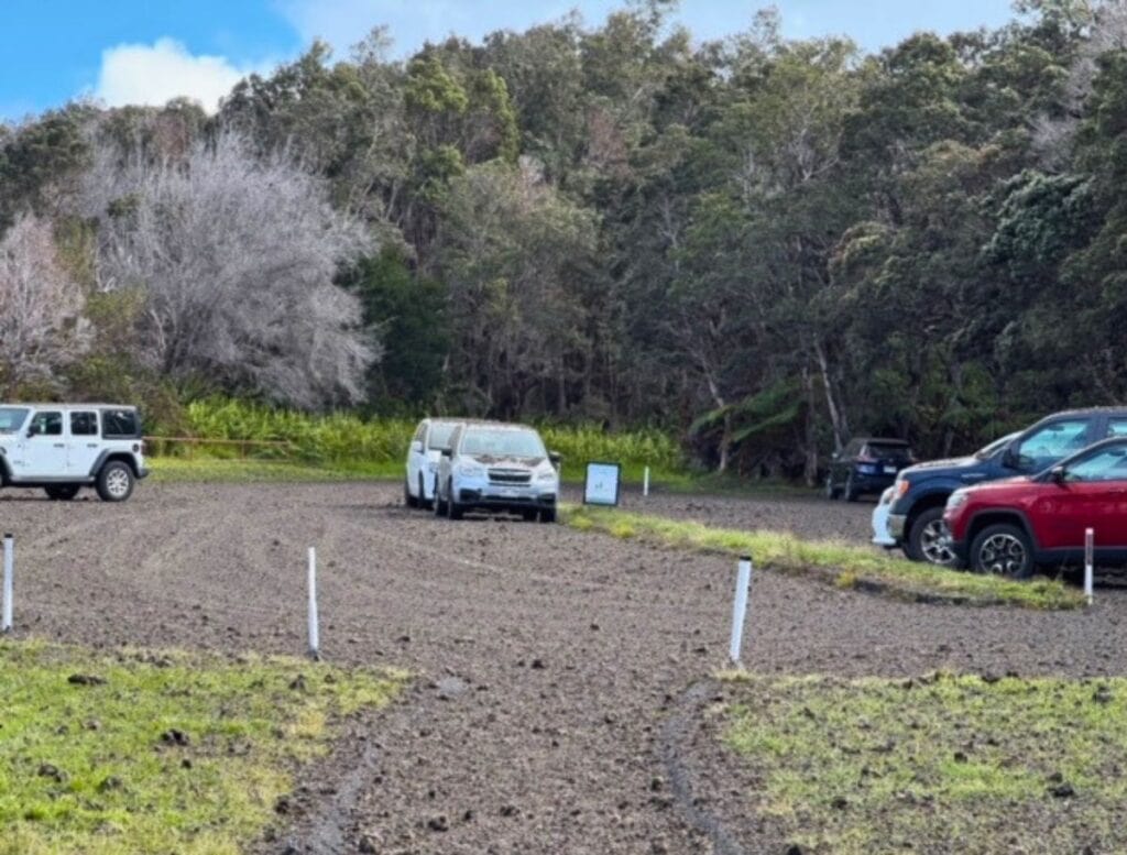 Several parked cars sit on a gravel lot covered in dark volcanic ash and small rocks with a forest in the background. The loose tephra spread across the ground highlights the risk of tephra damage rental car tires and paint when driving or parking in volcanic areas.
